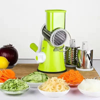 Manual rotary vegetable slicer with stainless steel drums next to sliced veggies on wooden cutting board.