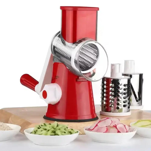 Manual rotary vegetable slicer in red with stainless steel drums, surrounded by sliced vegetables on a kitchen counter.